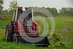 A red tractor sits in a rainy grass field with a forest in the background