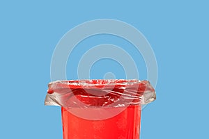 A red trash bin with a black bag inside stands on the surface close-up isolated on a blue background with a shadow at the bottom.