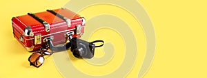 Red travel suitcase, sunglasses and a camera on a yellow background. Travel concept. Banner. Place for your text.