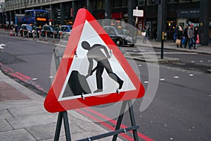 Red triangle warning roadworks sign placed at pavement, blurred city road with people, buses and taxis in background