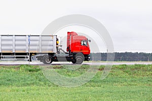 A red truck with a semi-trailer transports bulk cargo along the road in cloudy weather. Side view. Copy space for text