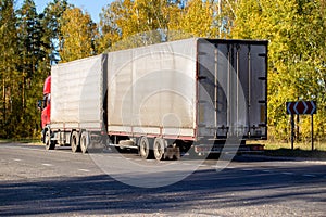 A red truck with a semi-trailer transports goods along a country highway against the backdrop of an autumn forest. Tonnage per photo