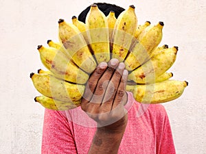 Red tshirt south Indian man covering his face with a cluster of bananas