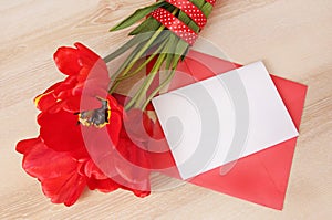 Red tulips bouquet with white paper card&envelope on wooden table.