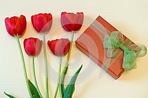 Red tulips and gift box with green bow on ight pastel background. Beautiful spring floral layout. Greeting card for women`s or