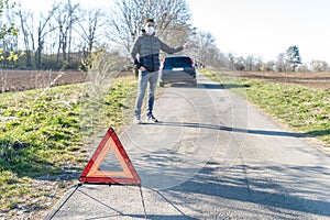 Red warning triangle on the road in front of a broken car. The young man is looking for help stopping vehicles moving