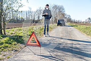 Red warning triangle on the road in front of a broken car. A young man using a telephone to order assistance service