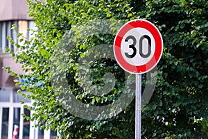 Red and white 30 speed limit sign with lush green tree in the background