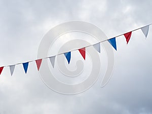 Red, white and blue festive bunting flags against sky background. Triangle shapes against cloudy sky.