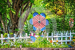 Red White and Blue Patriotic pinwheel flower in garden by white picket fence with trees and shubbery behind