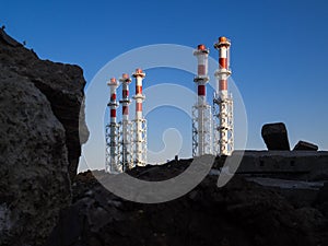 Red-and-white boiler room chimneys against a blue sky. Mountain of stones and debris and pipes boiler station