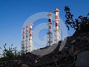 Red-and-white boiler room chimneys against a blue sky. Mountain of stones and debris and pipes boiler station