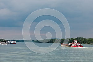 Red, white cargo ship sails along the wide part of River in cloudy weather, parked ships are in distance. selective focus. barge