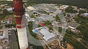 Red and white chimney-stalk above the industrial zone, hangars and other factory buildings surrounded by green forest