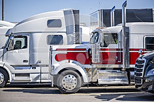 Red and white classic big rig semi truck with low profile cab and high exhaust pipes standing in row with another semi trucks and