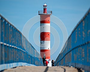Red and white lighthouse at the end of a blue walkway.Tourists gather