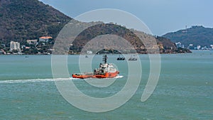 Red and white Offshore tug/Supply ship passes underway along the Vung Tau peninsula coastline