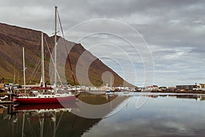 Red and white sailing boat reflected on the water, village, mountains, cloudy sky. Iceland