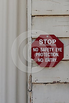 Red and white sign warning, Stop, No Safety Eye Protection, No Entry on the exterior of a weathered workplace building