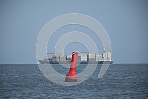 A red and white steel navigational floating buoy and a lrge containership in the blue sea water, drift with the sea wave, among