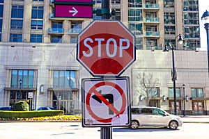 A red and white stop sign with a no left turn sign on a tall black pole with cars driving on the street and office buildings