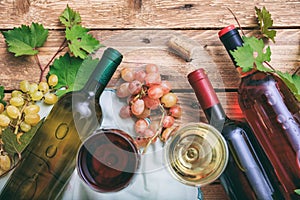 Red and white wine glasses and bottles on wooden background, copy space. Fresh grapes and grape leaves as decoration