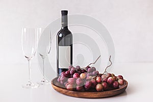 Red wine bottle, large burgundy fresh grapes on wooden dish, empty wine glasses on white background, mockup