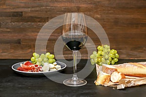 Red wine in glass, brie cheese, blue cheese, baguette, bread, grapes, jamon meat, salami on plate on black wooden table