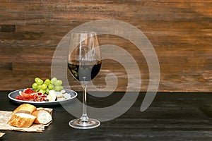 Red wine in glass, brie cheese, blue cheese, baguette, bread, grapes, jamon meat, salami on plate on black wooden table