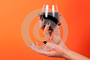 Female hand with black nails holding wine glass on orange background in studio