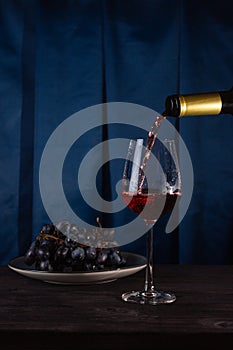 Red wine is poured from a bottle into a glass glass and grapes on a plate. Fabric blue background