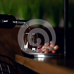 Red wine pouring from bottle into glass and bunch of ripe grapes on bar. Expensive alcoholic drink. Copy space. Soft