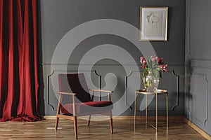 Red wooden armchair next to gold table with flowers in living room interior with poster