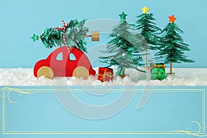 Red wooden car carrying a christmas tree over snow in front of blue background.