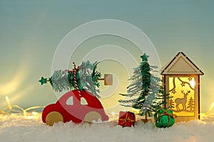 Red wooden car carrying a christmas tree over snow in front of blue background and golden garland lights.