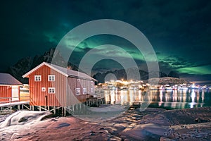 Red wooden house in fishing village with aurora borealis over Reine town in winter at night