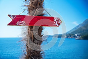 A red wooden sign with intricate patterns pointing towards the sea, hanging on a palm tree