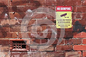 Red, yellow and black No Dumping, 24 Hours Video Surveillance, Violators Will Be Prosecuted warning sign on a brick wall