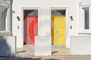 Red and yellow door side by side on white building facade, basking in sunlight.