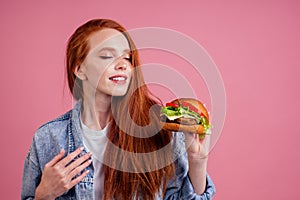 Redhaired ginger woman with freckles enjoying big burger cutlet and wearing demin american jeans jacket in studio pink photo