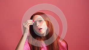 Redhaired ginger woman in pink studio background holding mascara apply on eyeleashes looks at camera like at mirror