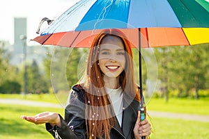 Redhaired ginger woman standing under colorful umbrella in summer park checking for rain rainbow