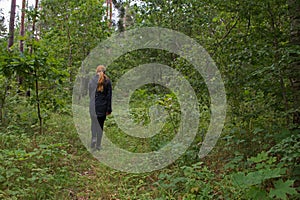 Redhaired girl in a black jacket and leggings is on the green grass of the forest among the trees