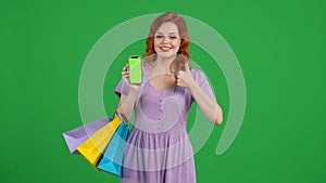 Redhaired woman with shopping bags holds a smartphone with a green screen and shows a thumbs up. Young woman in studio