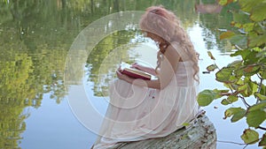 Redhead attractive caucasian girl reading the book in dark red cover on the bank of the lake in the summer forest. Fairy