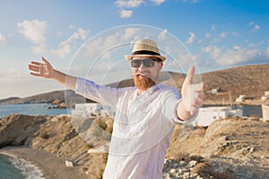 Redhead bearded hipster traveler man with open hands in hat and sunglasses smiling against the blue sea and sky background