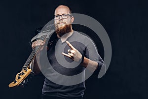 Redhead bearded male musician wearing glasses dressed in a gray t-shirt holds electric guitar and show rock and roll