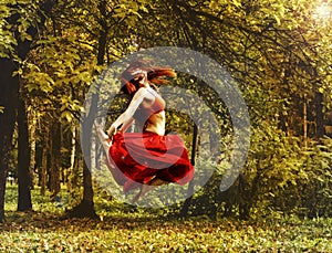 Redhead beautiful woman in red dress dancing in a autumn forest.