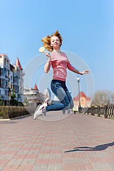 Redhead beautiful young woman jumping high in air over blue sky holding colorful lollipop. Pretty girl having fun outdoors.