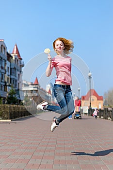 Redhead beautiful young woman jumping high in air over blue sky holding colorful lollipop. Pretty girl having fun outdoors.